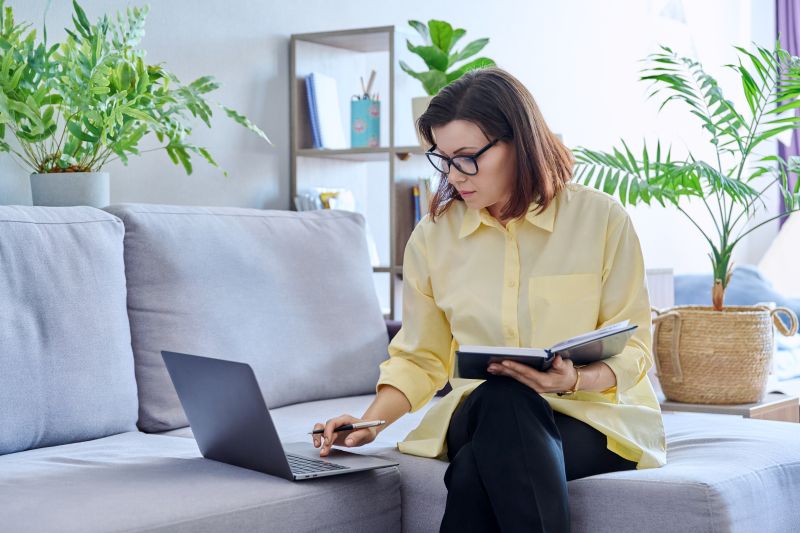 woman on couch at home doing research on laptop with book