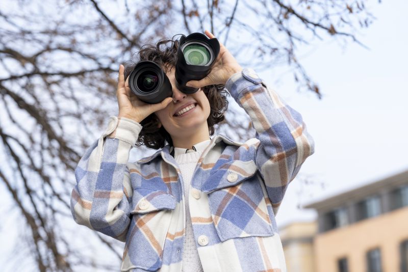 Young brunette curly hair woman fooling around with two photography lenses. She is putting the lenses over her eyes.